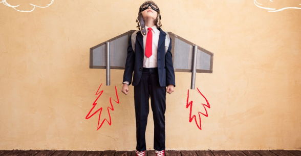 A young boy in coat and time wears atique pilot goggles while standing in front of wings painted on a wall